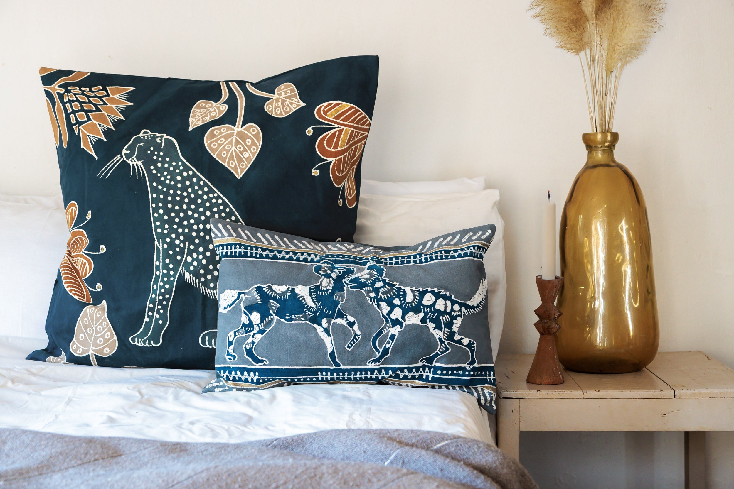 Decorative pillows with animal patterns - wild dog and cheetah on a bed next to a wooden side table with a gold vase.
