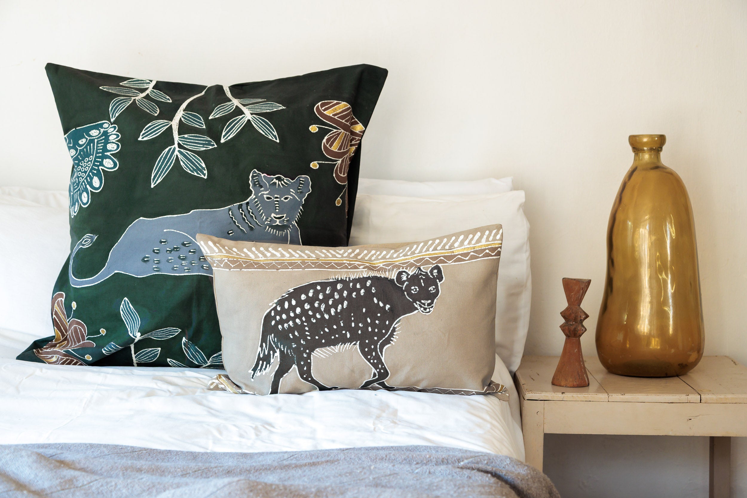 Decorative pillows with animal prints Lion and Hyena on a bed next to a nightstand with a gold vase.