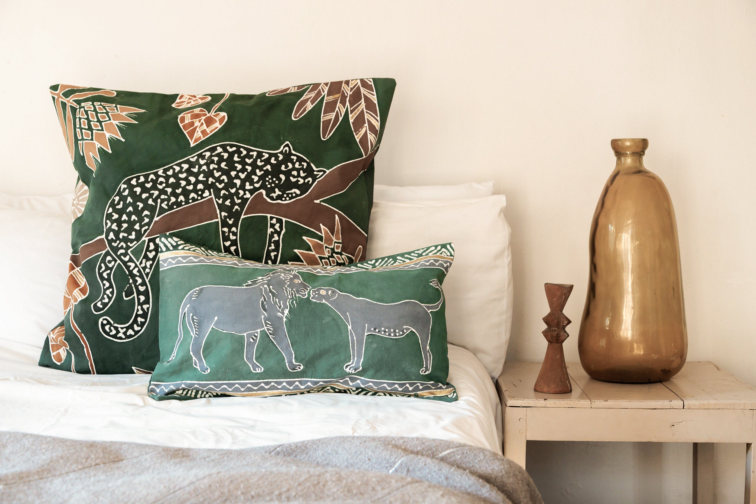 Decorative pillows with animal patterns Lion and Leopard on a bed next to a wooden side table with a vase.