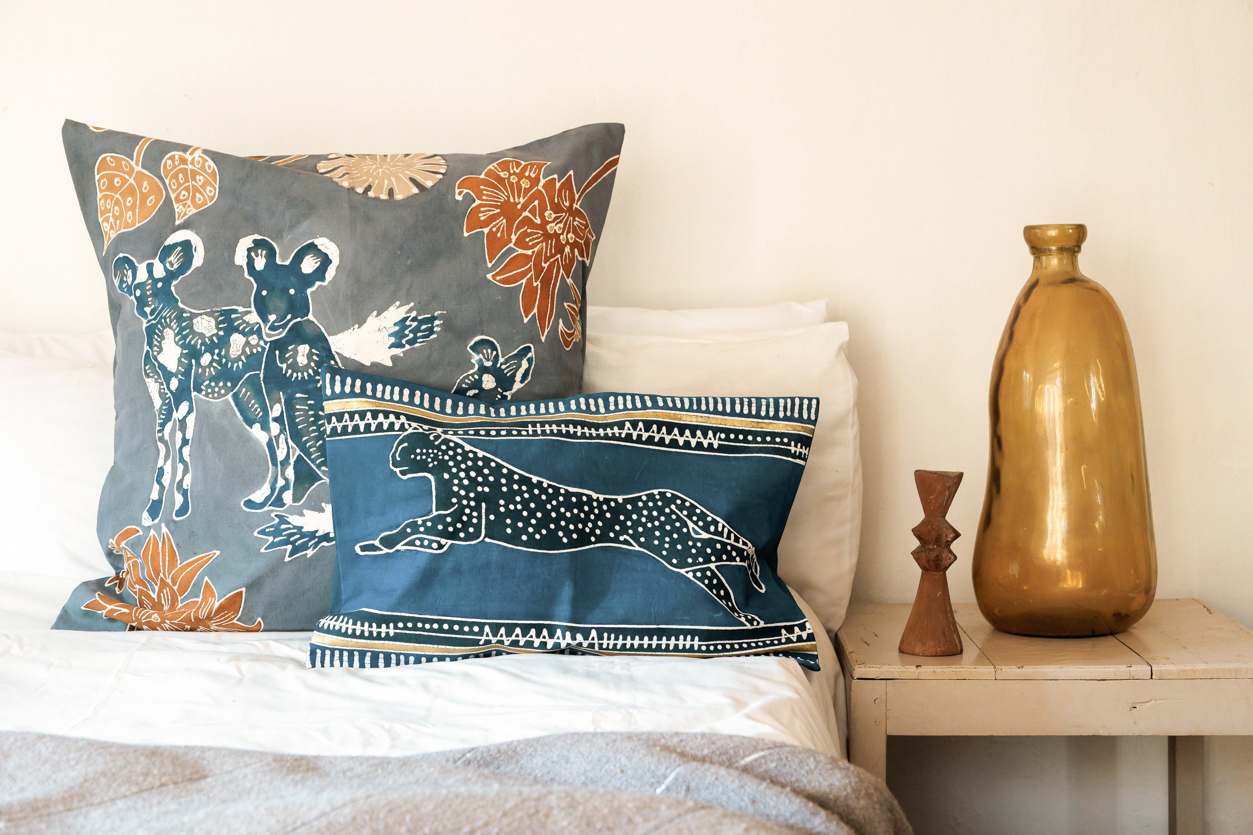 Decorative pillows with animal patterns - Wild Dogs and Cheetah on a bed next to a wooden nightstand with a gold vase.